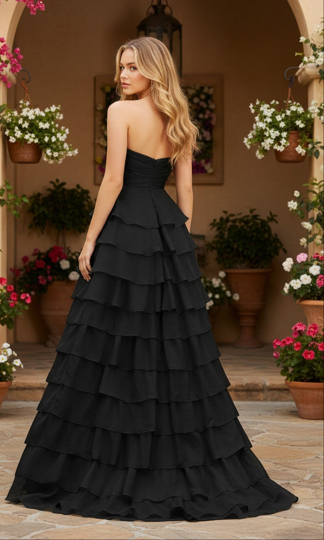 A woman wearing the PromGirl Exclusive 250233B long prom dress with a tiered ruffle skirt stands outdoors on a stone path, surrounded by potted flowers and hanging plants.